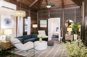 A cozy room with a blue sofa, a white coffee table, and a ceiling fan. There is a bed with a white headboard and a nightstand with a lamp.