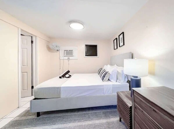 A hotel room with a bed, bedside table, lamp, air conditioning unit, and a door.