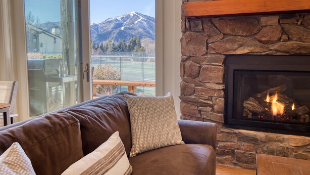 Ein gemütliches Wohnzimmer mit Kamin aus Stein, einer Ledersofa und Bergblick durch eine Glastür.