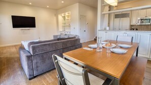 Ein modernes Wohnzimmer mit einem grauen Sofa, einem Holzesstisch für vier Personen und einer Küchenzeile mit weißen Schränken und einem eingebauten Backofen.