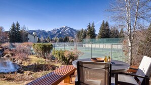Eine Holzterrasse mit Tisch und Stühlen mit Blick auf einen Tennisplatz und ein Gebirge.