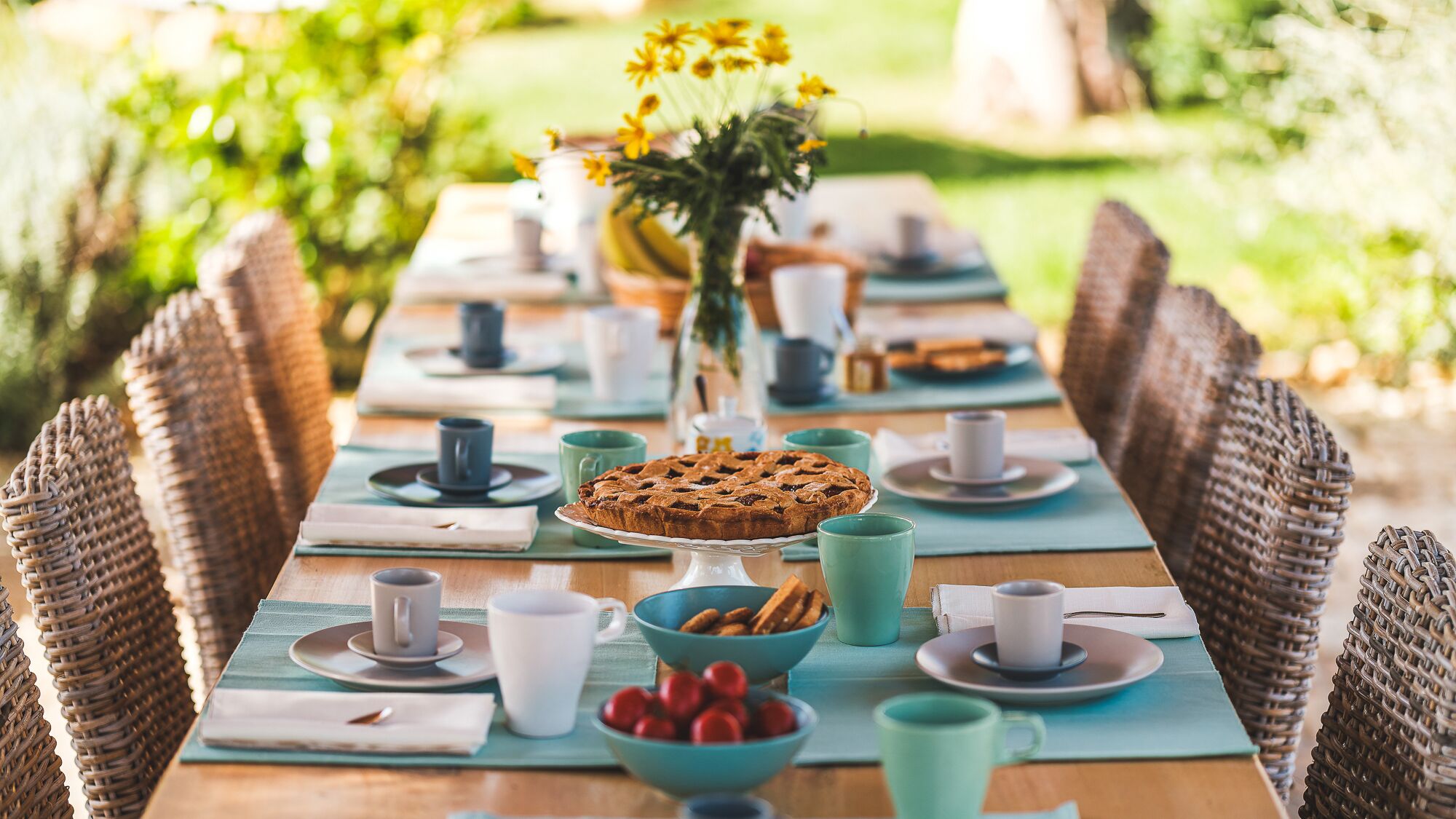 Une longue table à manger, dressée avec des assiettes, des tasses et un gâteau au centre, entourée de chaises en osier.