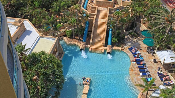Vue aérienne d’un parc aquatique avec une grande toboggan aquatique au centre, des palmiers tout autour et un espace piscine avec des chaises longues.