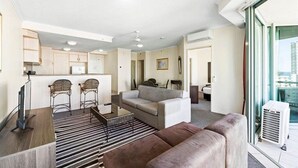 Un salon moderne avec un canapé, une table basse et un téléviseur. Il y a un coin cuisine équipé de placards et d’un réfrigérateur. La pièce est dotée de grandes fenêtres et offre une vue sur la ville.