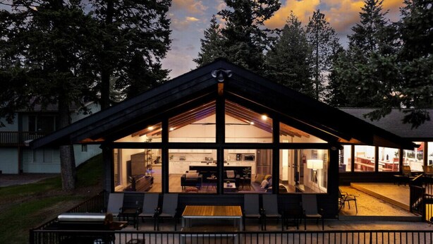 A modern house with a large glass front, a deck with seating, and a visible kitchen area.