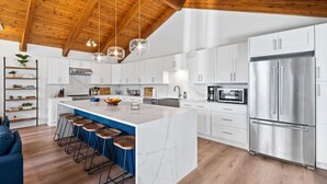 A modern kitchen with a large island, white cabinetry, stainless steel appliances, and wooden ceiling beams.