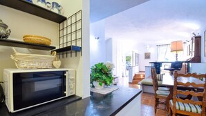 Een keuken met een magnetron, planken met keukengerei, een eethoek met een houten stoel en uitzicht op de woonkamer.