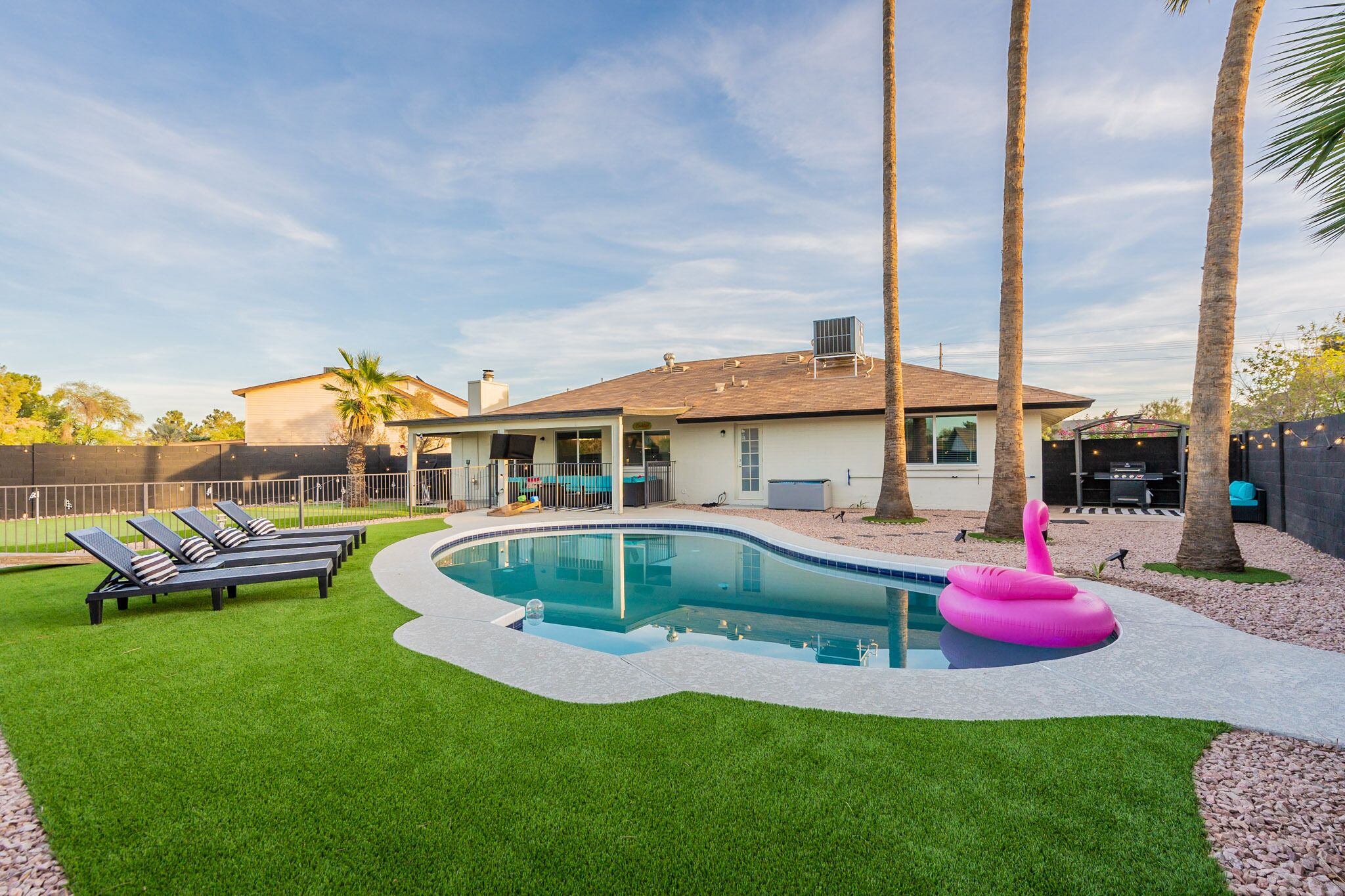 A swimming pool with a curved edge, surrounded by artificial grass, palm trees, and a house with a covered patio.
