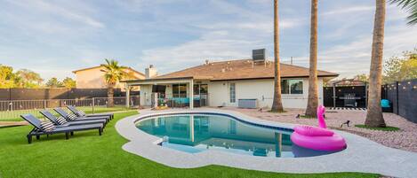 A swimming pool with a curved edge, surrounded by artificial grass, palm trees, and a house with a covered patio.