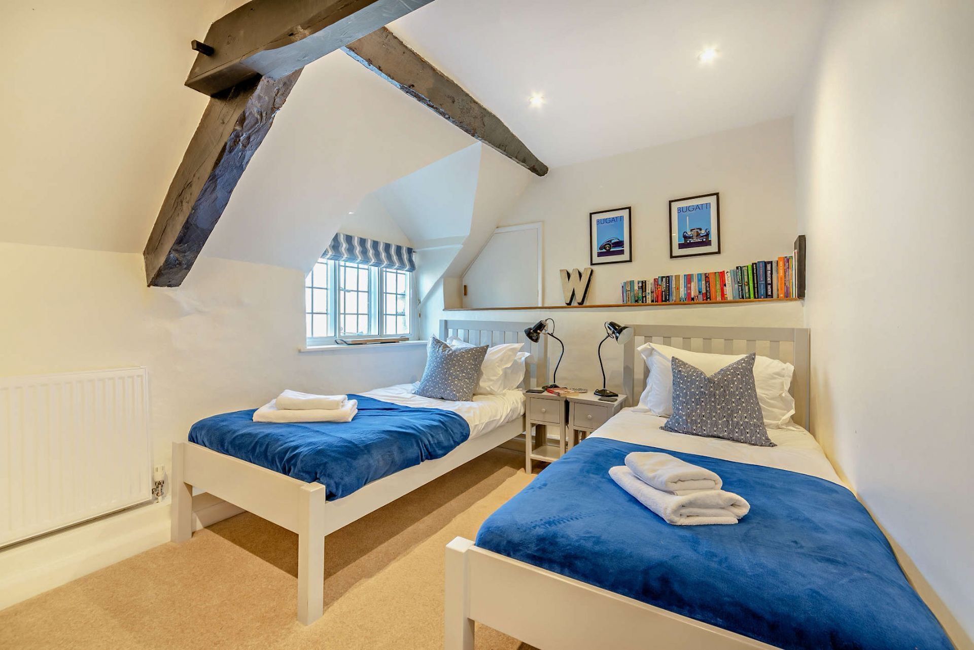 Een slaapkamer met twee bedden, een raam, een radiator, een boekenplank en een plafond met zichtbare balken.