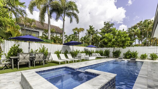 Zwembad met betegelde rand, omringd door ligbedden en parasols, met palmbomen en een witte hek in de achtergrond.