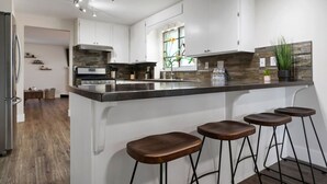 A modern kitchen with white cabinets, a large island with stools, a built-in oven, and a window with a view. - Glacier Getaway! Huge Home! Sleeps 14! (Columbia Falls)