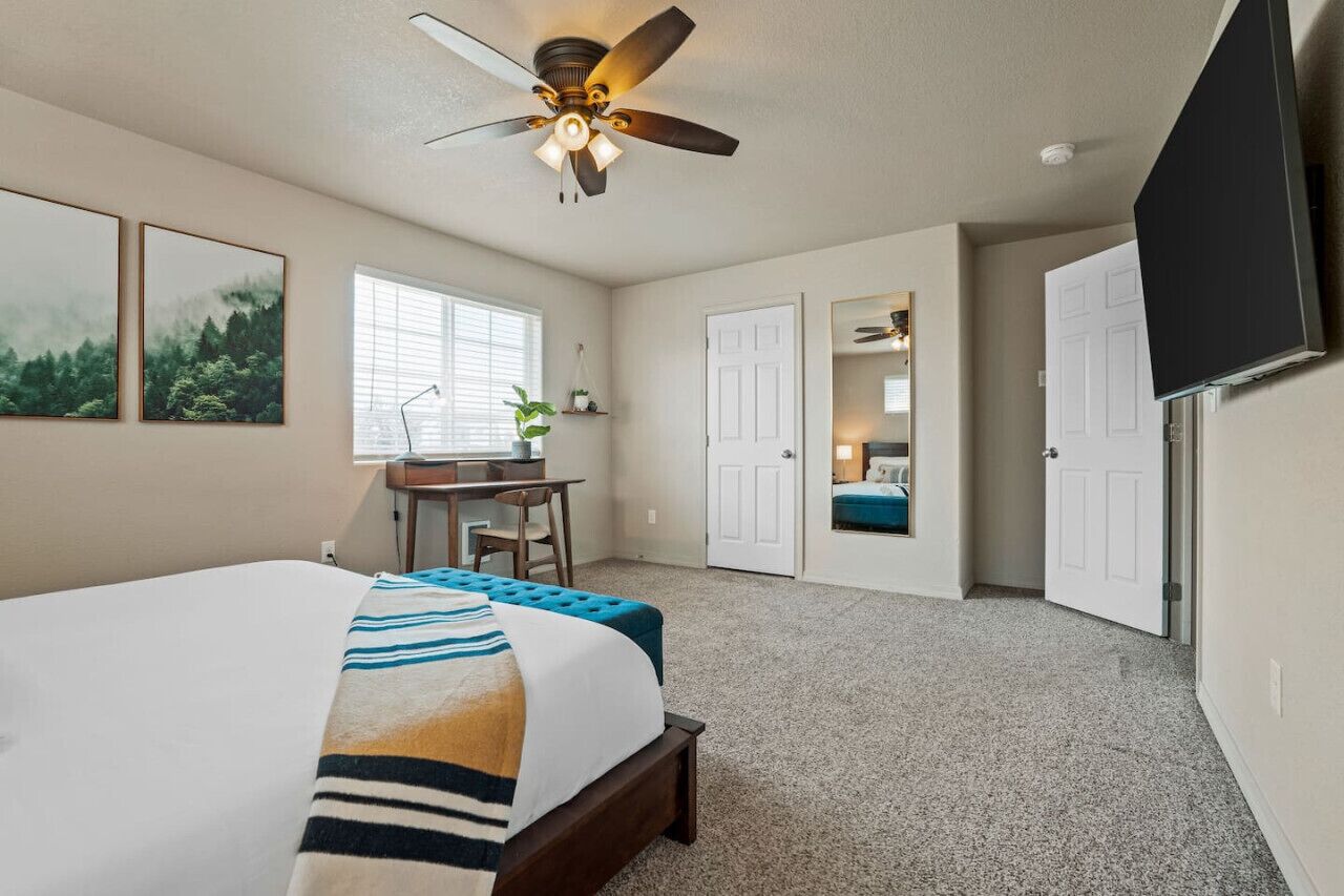 A bedroom with a bed, a desk, a chair, a ceiling fan, and a TV mounted on the wall.