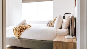 A modern bedroom with a large window, a bed with white linens, multiple pillows, a throw blanket, and a bedside table with a lamp. - Wombat Hollow - Holiday Houses Tasmania (Swansea)