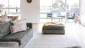 A modern living room with a grey sofa, a small ottoman, and a large window offering a view of the outdoors. - Wombat Hollow - Holiday Houses Tasmania (Swansea)
