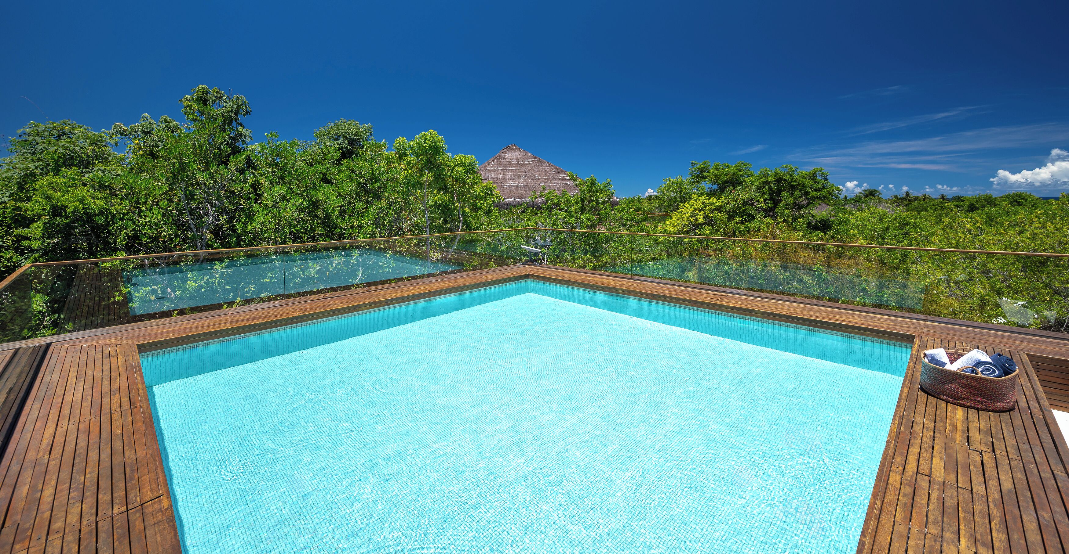 A clear blue infinity pool with wooden deck, surrounded by lush greenery and a thatched-roof structure in the background.