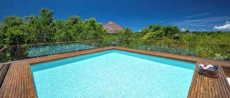 A clear blue infinity pool with wooden deck, surrounded by lush greenery and a thatched-roof structure in the background.
