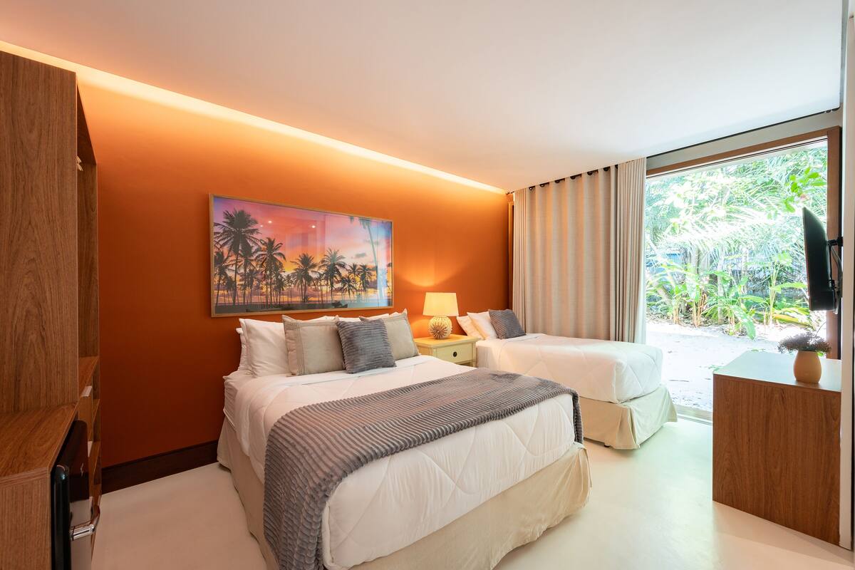 A hotel room with two beds, a painting of palm trees, a nightstand with a lamp, and a view of greenery outside.