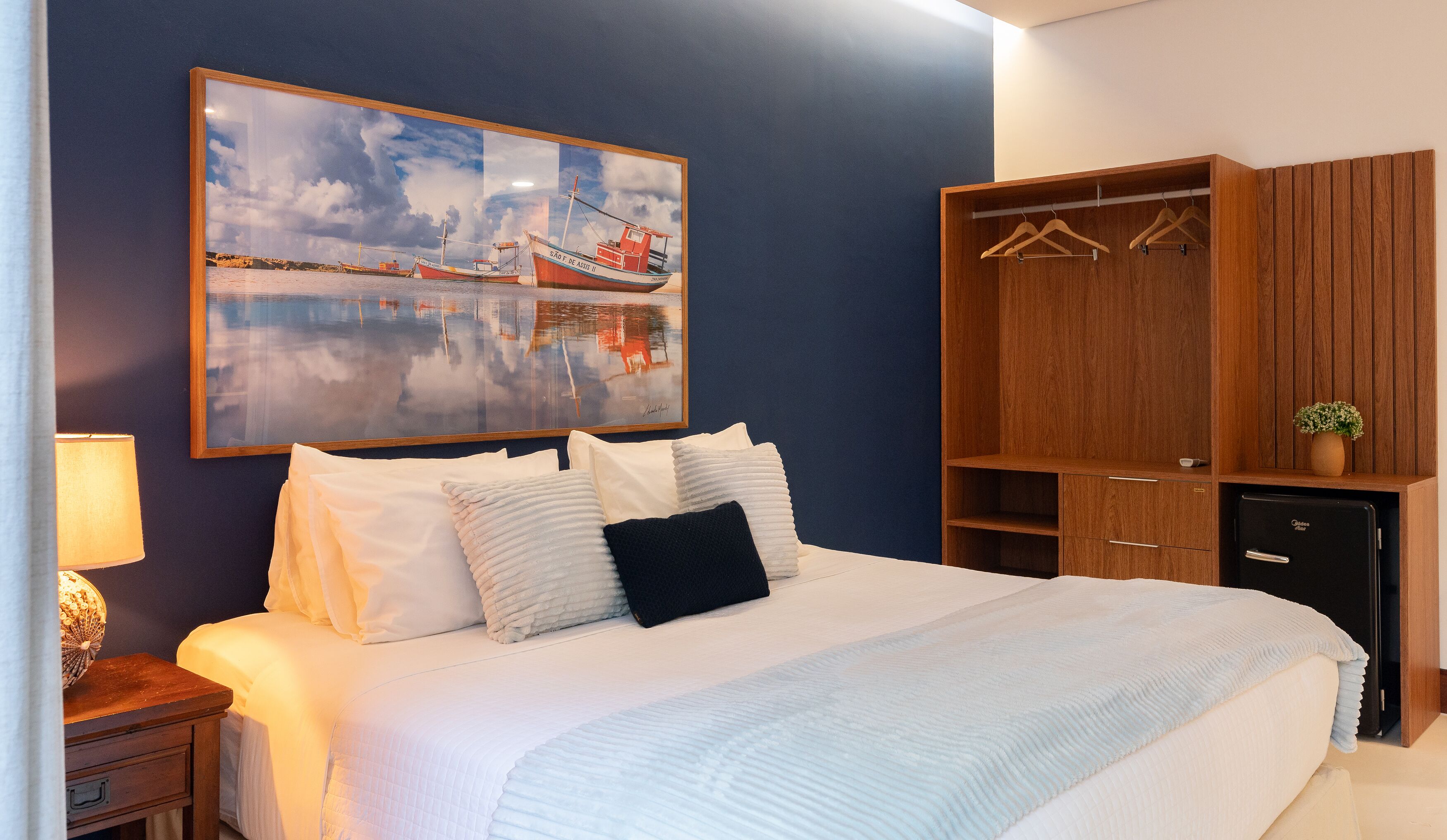 A neatly made bed with white linens and a blue pillow, a wooden wardrobe with hangers, a small potted plant, and a framed painting of boats on a wall.