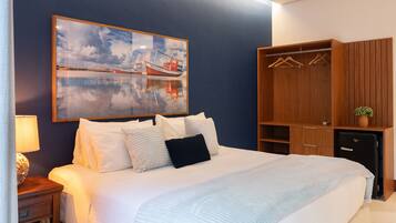 A neatly made bed with white linens and a blue pillow, a wooden wardrobe with hangers, a small potted plant, and a framed painting of boats on a wall.