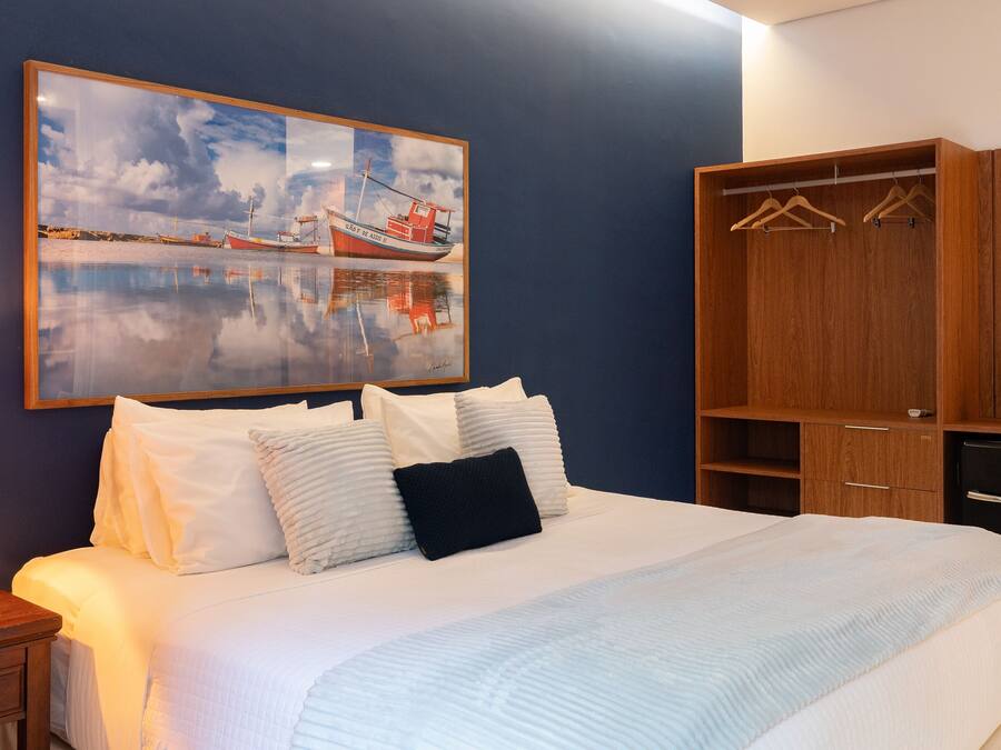 A neatly made bed with white linens and a blue pillow, a wooden wardrobe with hangers, a small potted plant, and a framed painting of boats on a wall.