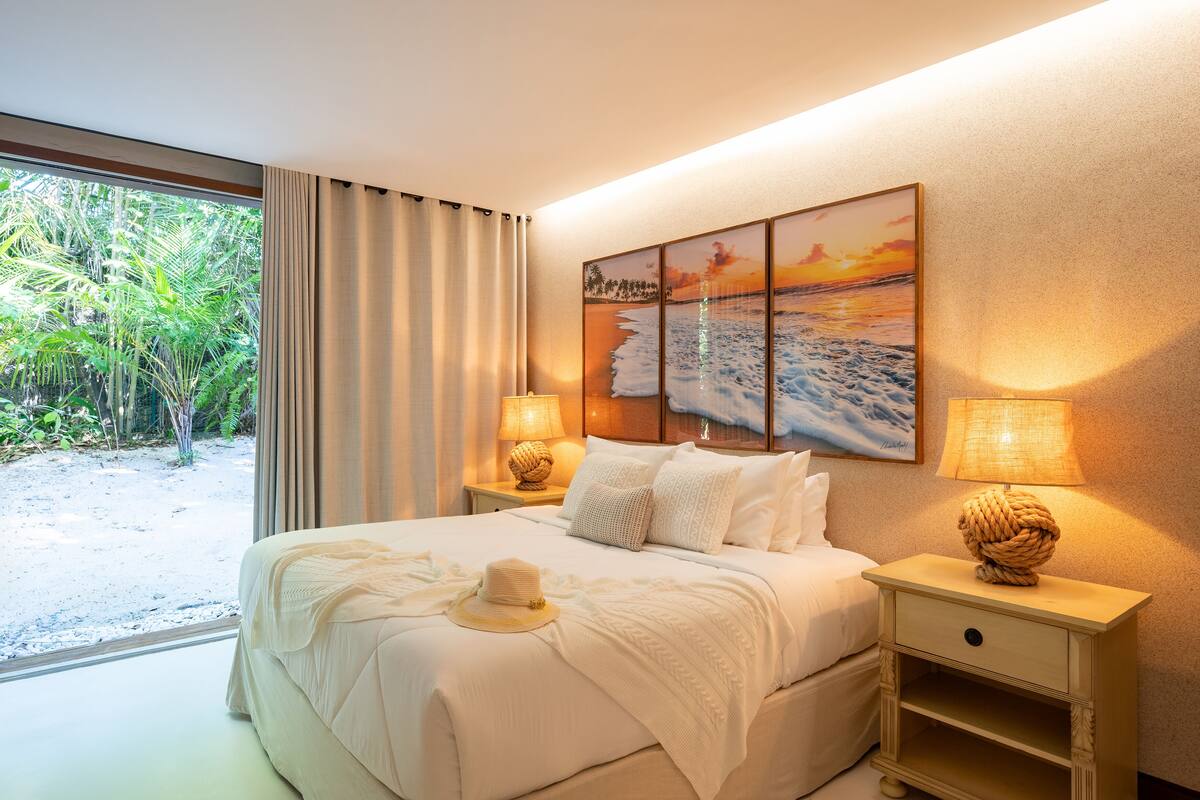 A hotel room with a large bed, two bedside lamps, a wall-mounted painting, and a view of greenery outside.