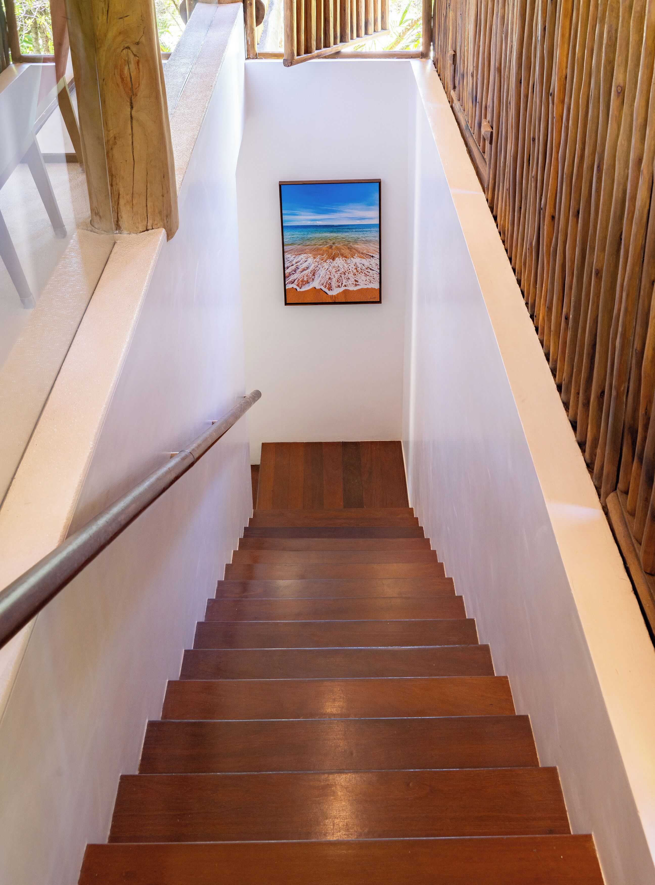 Una escalera de madera con barandilla, paredes blancas y un cuadro enmarcado con una escena de playa.