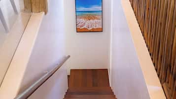 Una escalera de madera con barandilla, paredes blancas y un cuadro enmarcado con una escena de playa.