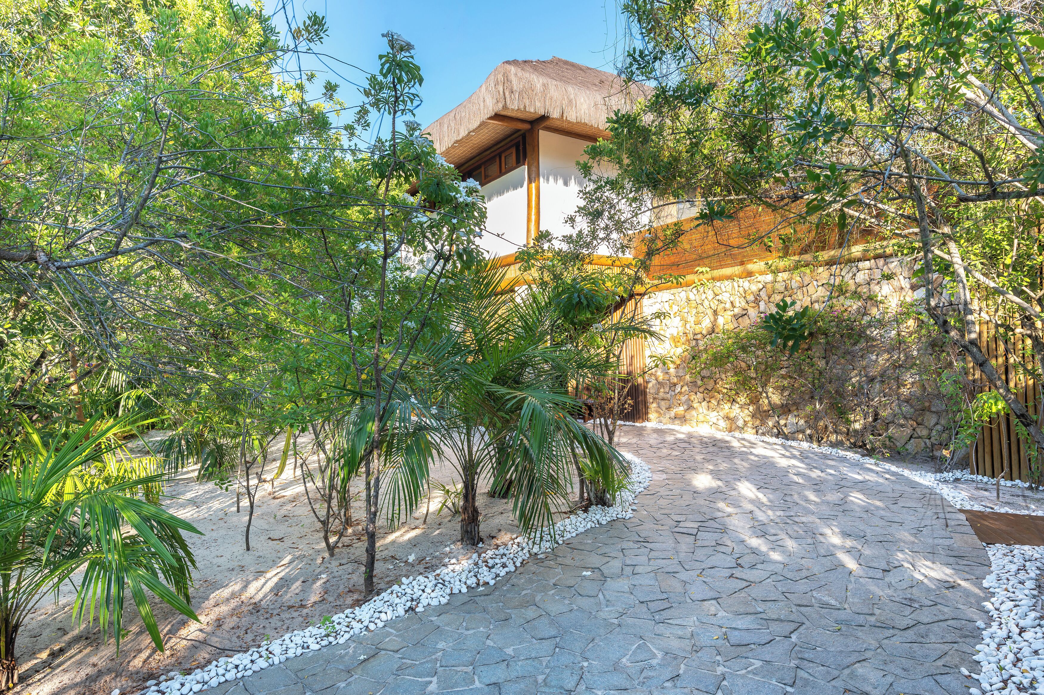 A stone pathway leading to a building with a thatched roof, surrounded by lush greenery and white pebbles.