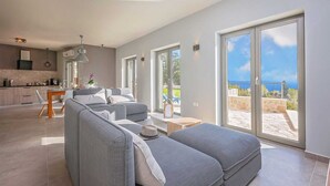 A modern living room with a grey sofa, a dining area with a table and chairs, a kitchen in the background, and large sliding glass doors leading to a balcony with a view of the sea. - Villa Soleil in peaceful location with a sea view (Mali Kosi)