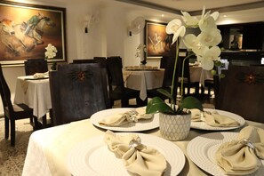 Uma sala de jantar com mesas postas para quatro pessoas, cada uma com uma toalha de tecido dobrada e uma orquídea branca num vaso estampado.