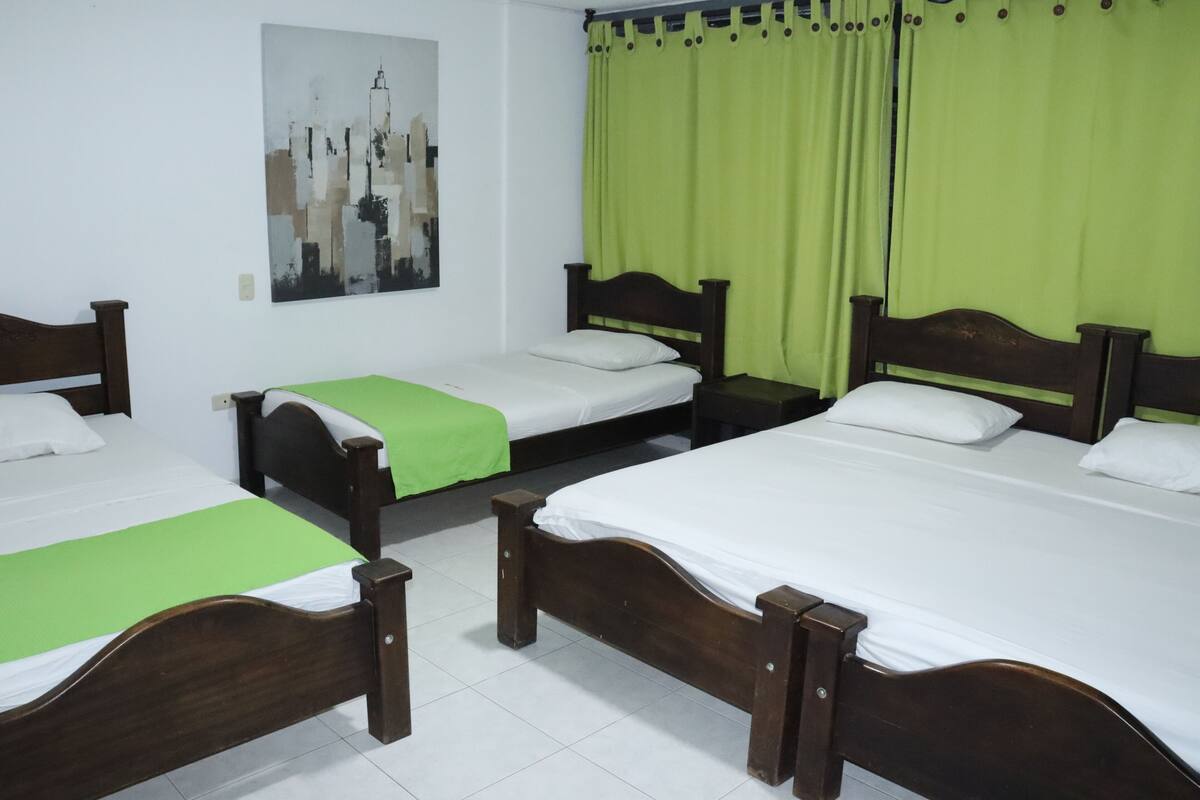 Una habitación con tres camas de madera, cada una con ropa blanca y colchas verdes. Hay una cortina verde en un lateral de la habitación.