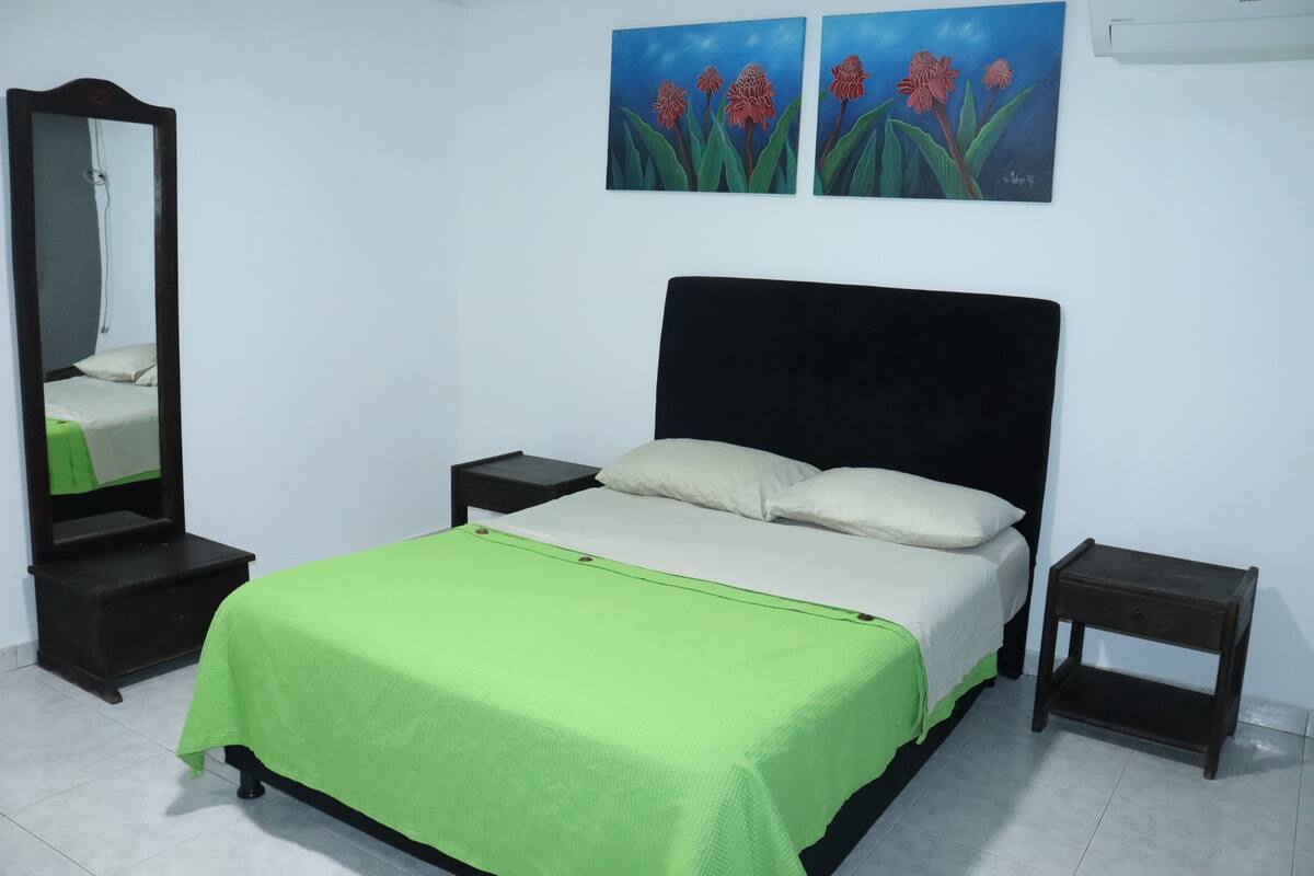 Un dormitorio con cama, mesita de noche, cómoda, espejo y dos cuadros colgados en la pared.