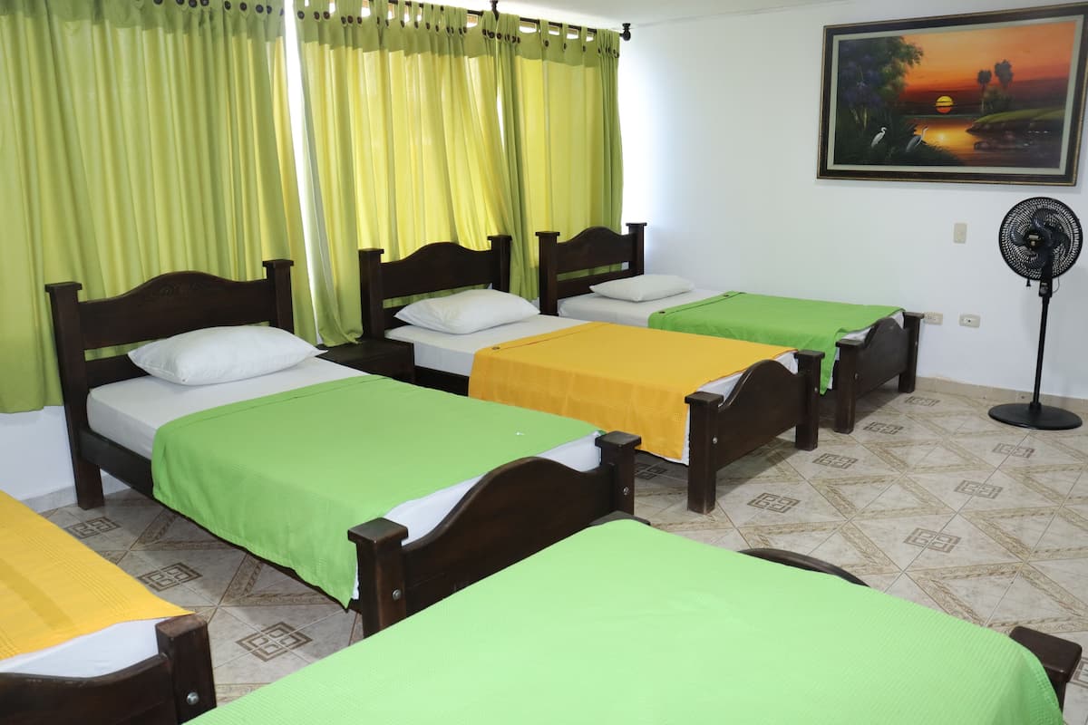 Four single beds with wooden headboards and green bedspreads, a large window, and a painting of a sunset adorn this inviting room.