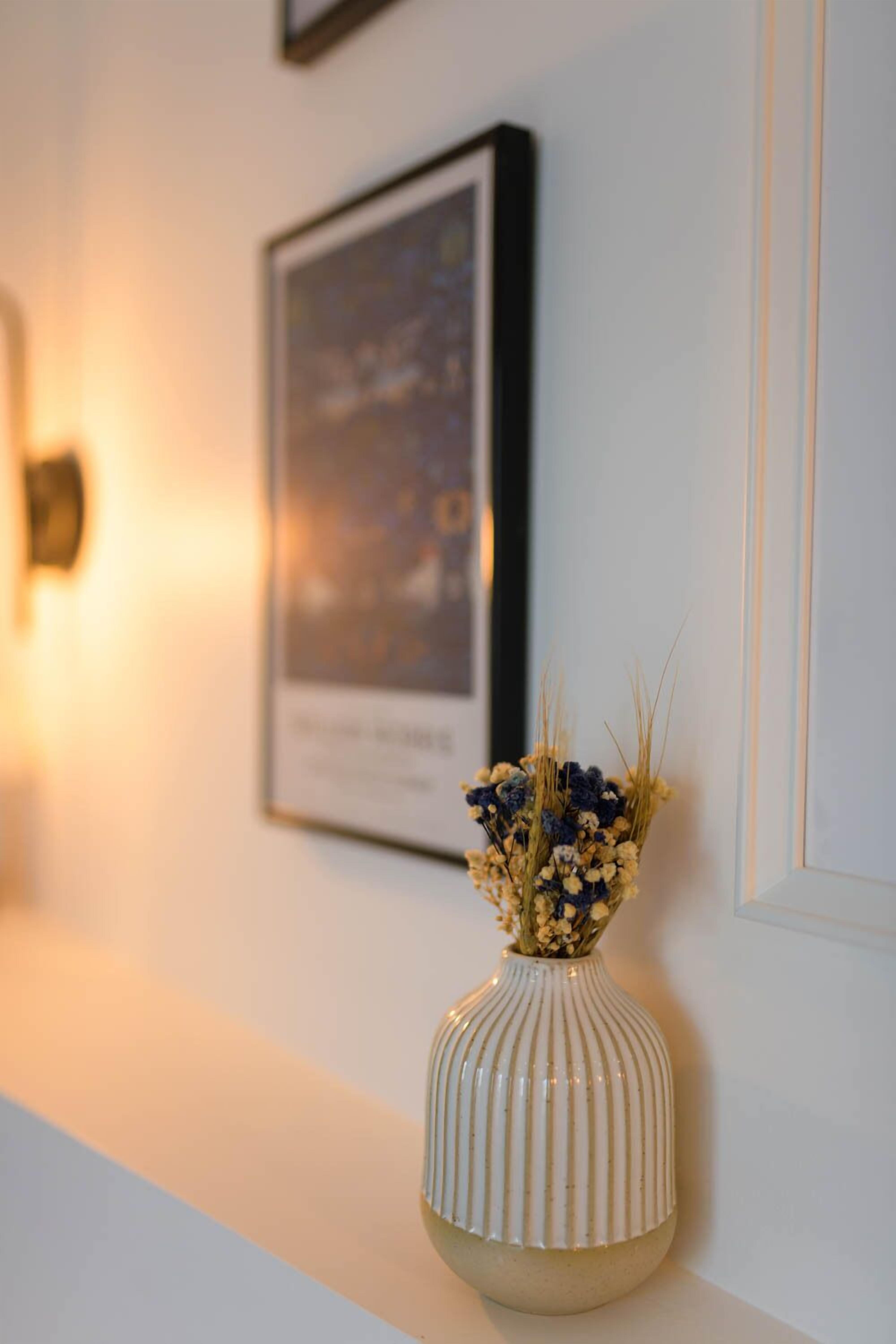 A white vase with a ribbed design on a white surface, a framed picture on the wall, and a small wall-mounted light.