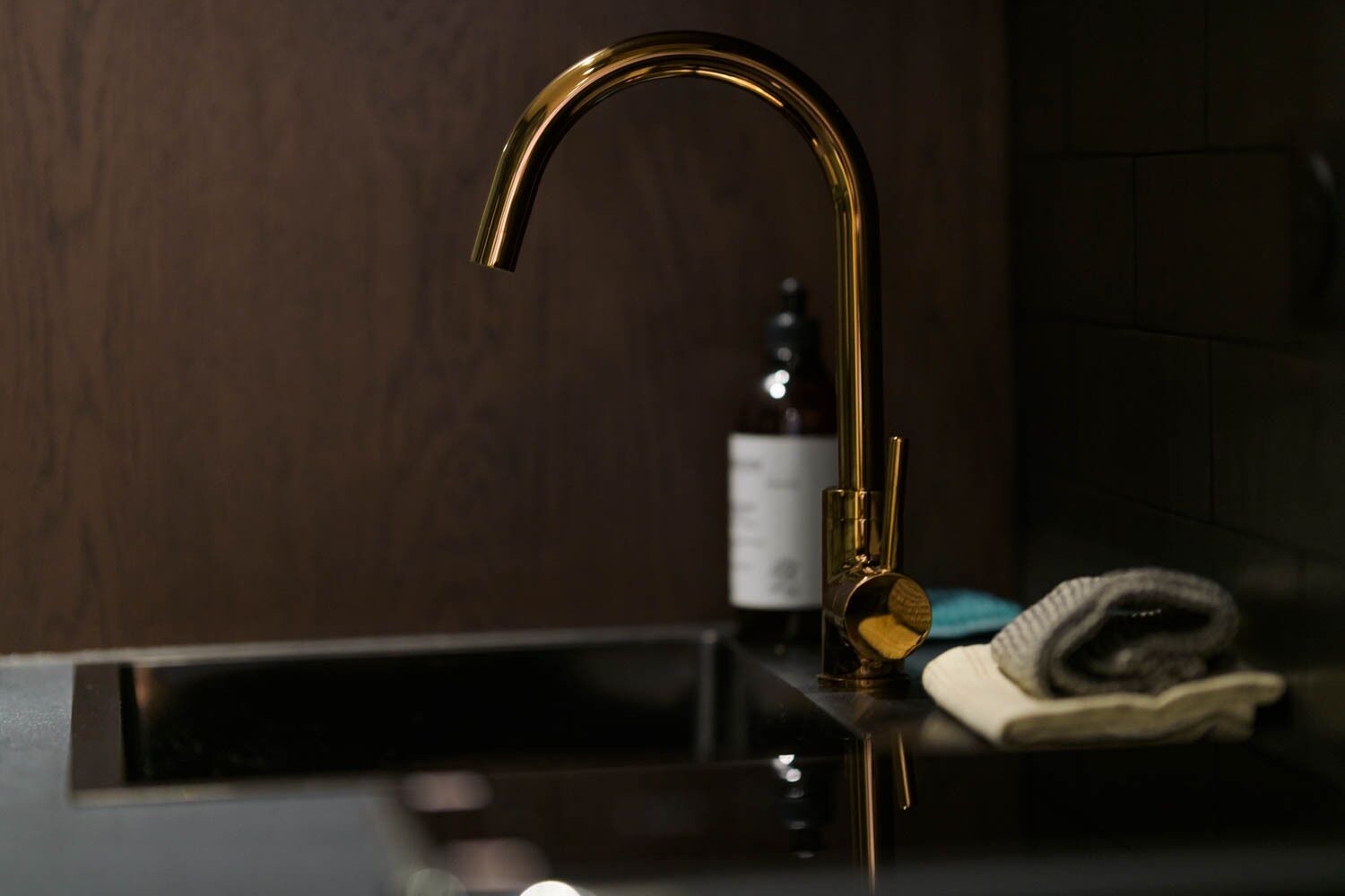 A modern bathroom with a gold faucet, a bottle of liquid, and a towel on the counter.