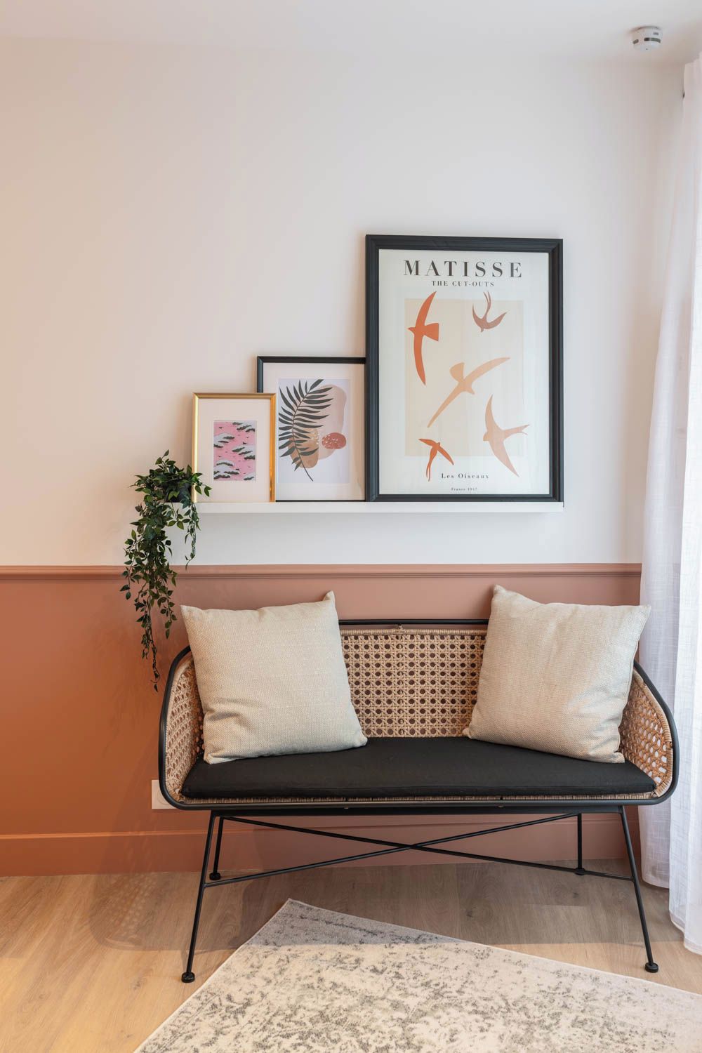 Een kamer met een zwarte bank met kussens, een geweven bankje en een lijstwerk met Matisse's 'The Cut-Outs' aan de muur.