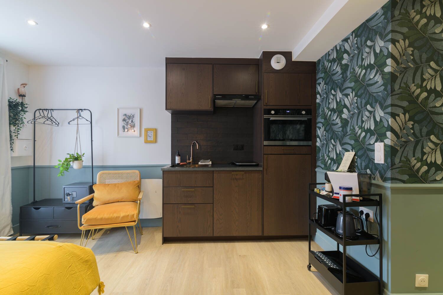 A compact kitchen with dark wood cabinets, a black countertop, and a built-in oven. There is a chair with a yellow cushion and a small side table with a plant.