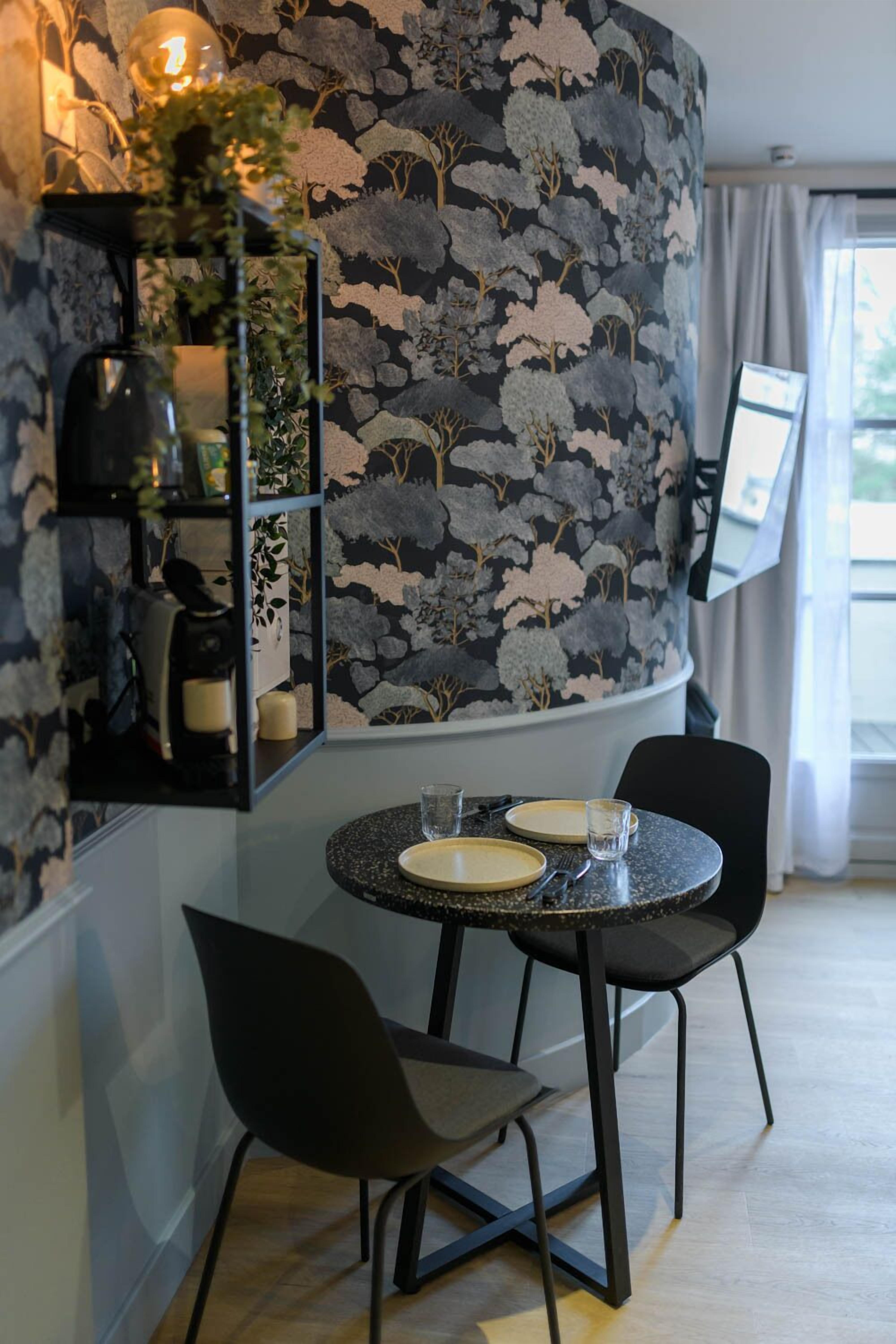 Un coin repas comprenant une table ronde, deux chaises, un mur orné d’un motif floral, une étagère avec des objets décoratifs et une fenêtre avec des rideaux.