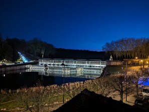 A nighttime view of a bridge over a reflective water body, surrounded by trees and a stone wall with inscriptions. - Le Logis d'Origine. (Marly-le-Roi)
