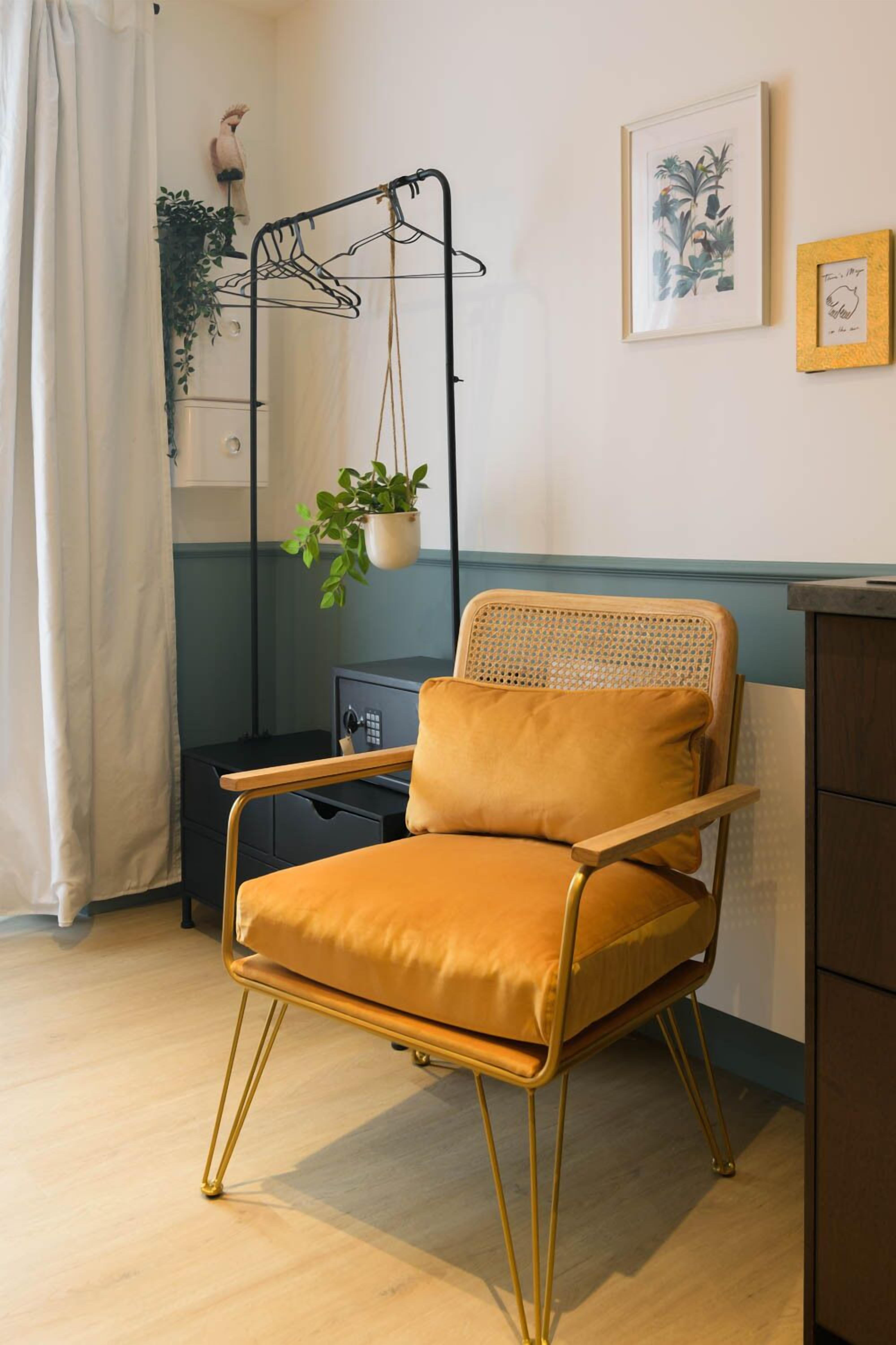 A room with a mustard-colored chair, a black metal drying rack, a potted plant, and a framed botanical print on the wall.