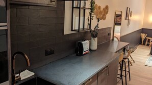 A modern kitchen with dark wood cabinets, a stainless steel sink, and a built-in oven. There is a small island with stools and a plant. - Le Logis d'Origine. (Marly-le-Roi)