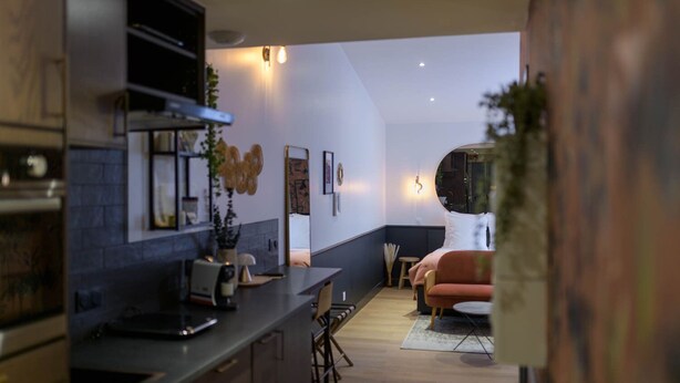 A modern kitchen with dark countertops, a built-in oven, and a dining area with a wooden table and chairs. - Le Logis d'Origine. (Marly-le-Roi)