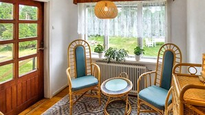 A room with a wooden door, a window with lace curtains, two wicker chairs with blue cushions, a small round table, and a radiator.