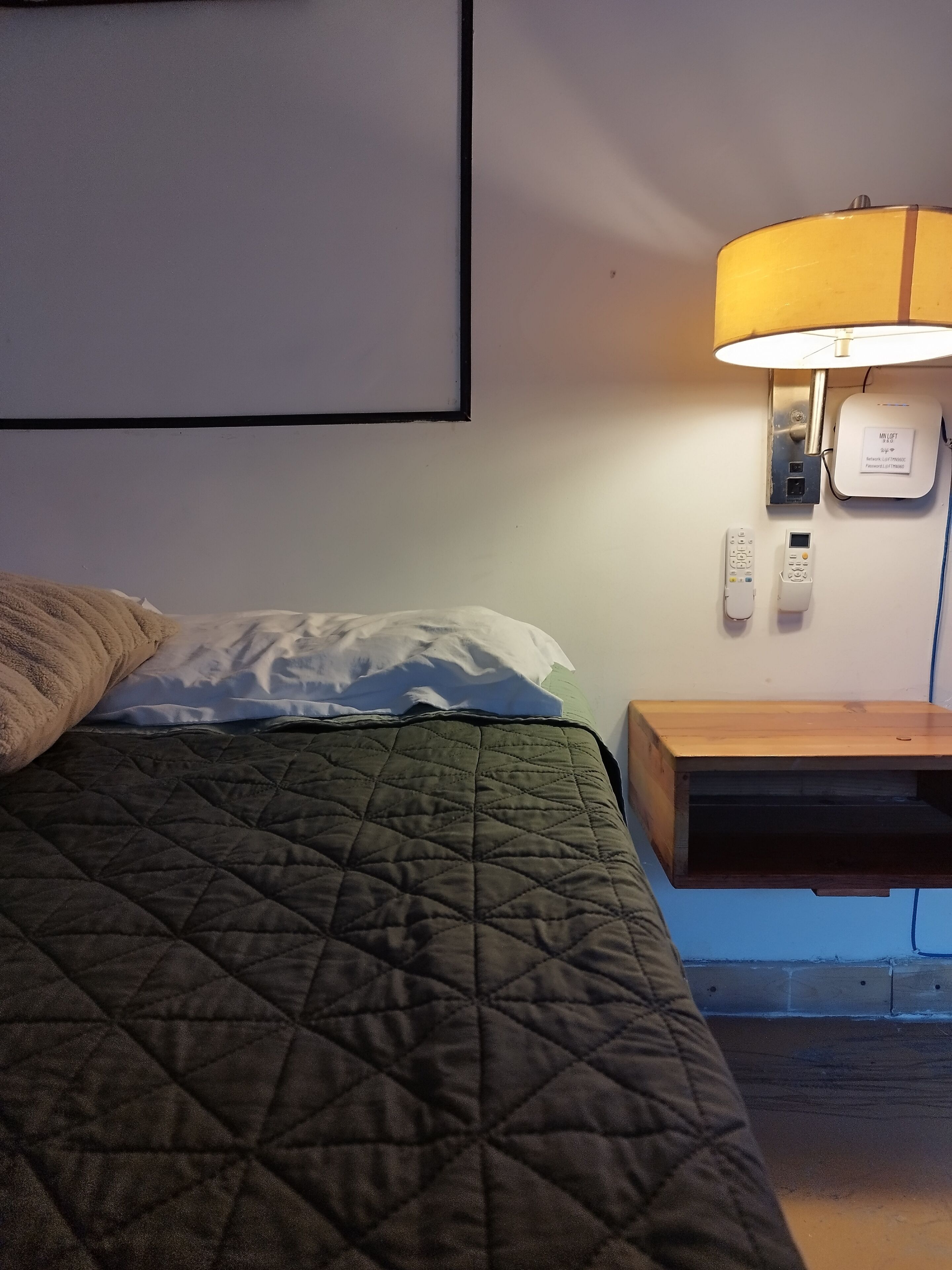 A bed with a green quilt, a wooden nightstand, a lamp, and a wall-mounted control panel.