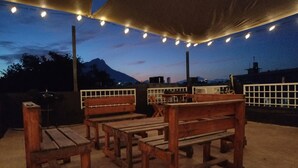 An outdoor seating area with wooden benches and tables, string lights, and a view of a mountain in the background. - DGM Lofts MTY C (Monterrey)