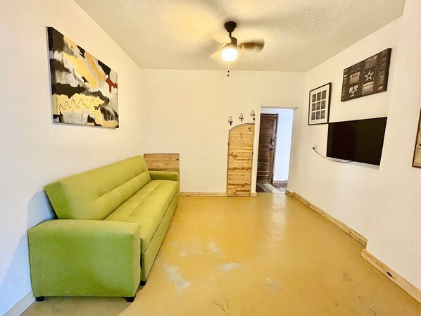 Una habitación con un sofá verde, un ventilador de techo, una puerta de madera, un televisor montado en la pared y un cuadro enmarcado.