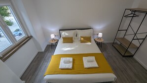 A neatly made bed with a yellow blanket and two pillows, two bedside lamps, a wooden shelf unit, and a window with a view of a car outside.