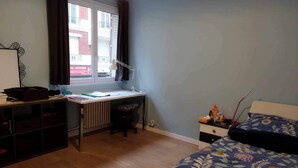 A bedroom with a wooden floor, a desk with a chair, a bed with a floral bedspread, and a window with curtains. - Paris, à 6mn de St Lazare, au Pied de la Gare, F3 Neuf, 7 Couchages max (Asnières sur Seine)