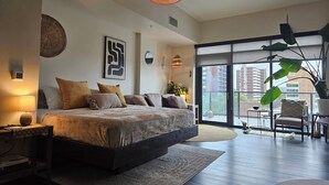 A modern bedroom with a large bed, a nightstand, a lamp, and a view of the city through large windows.